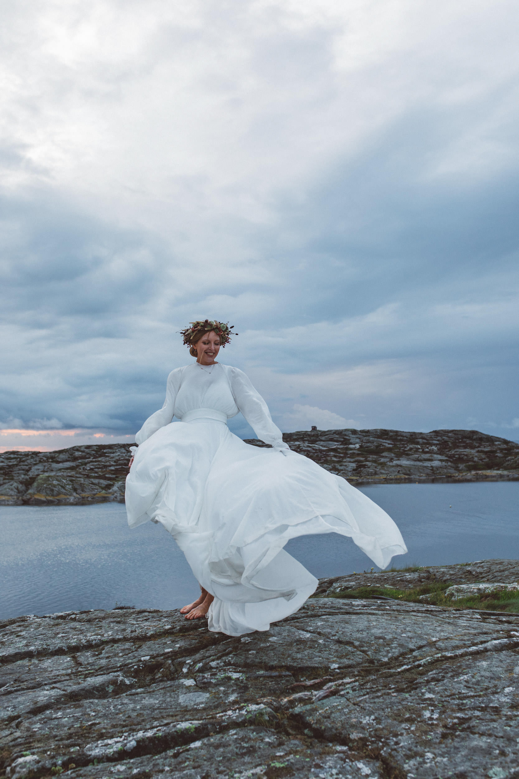Glad kvinna i vit brudklänning och blomsterkrans i håret står på en klippa på västkusten med dramatisk himmel och blåst i kjolen