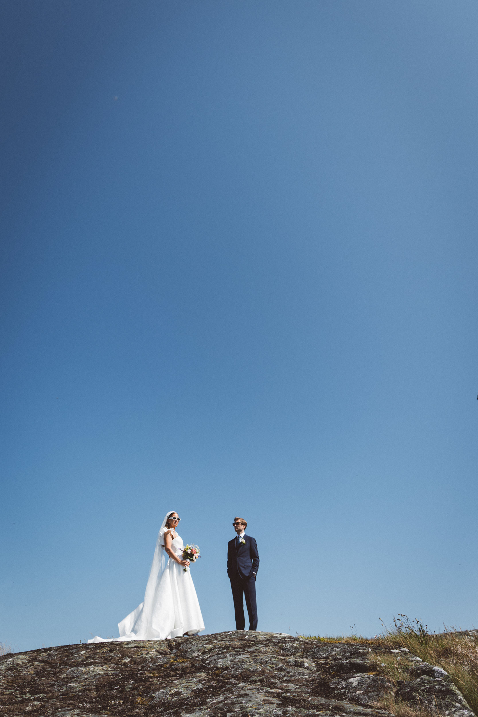 Brudpar med solglasögon står på en klippa under klarblå himmel